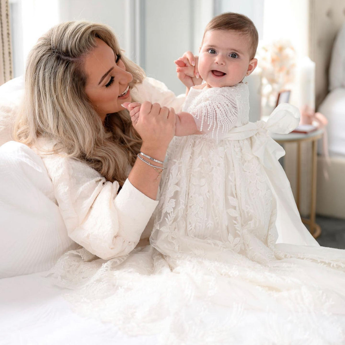 Mother holding her baby girl dressed in the My Blessing Elora ivory christening gown adorned with sequins, glass seed beads, and Swarovski crystal beadwork.