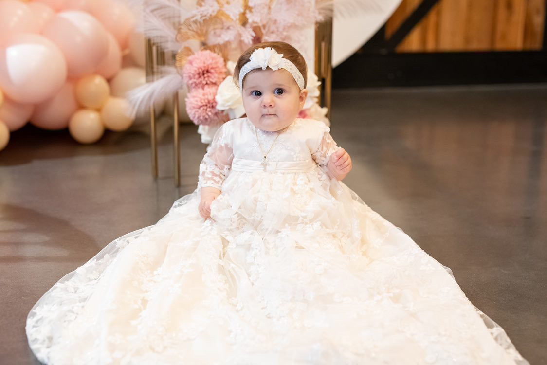 Baby girl sitting at the reception in her full‑length Christening gown, the floral lace overlay glowing beautifully in the ambient lighting.