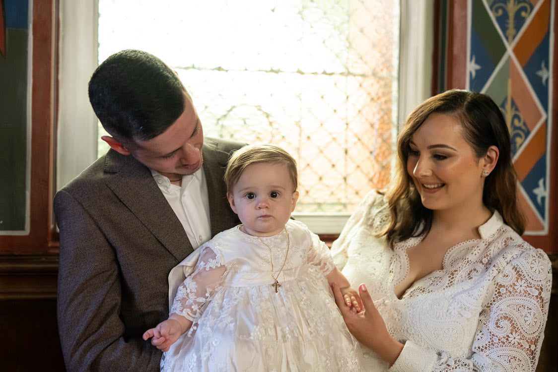 Baby girl wearing the My Blessing Faith christening gown being held by mum and dad inside a softly lit church, the European lace and glass seed beads catching the light beautifully.