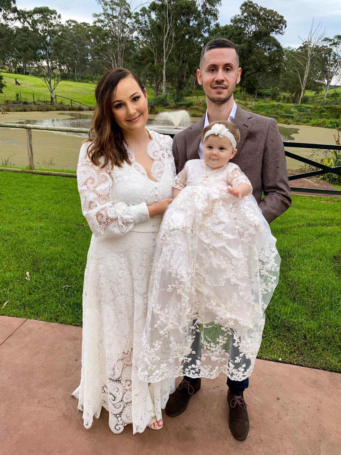 Mum and dad embracing their baby girl dressed in the My Blessing Faith christening gown, the elegant lace and beadwork enhancing the sacred moment.