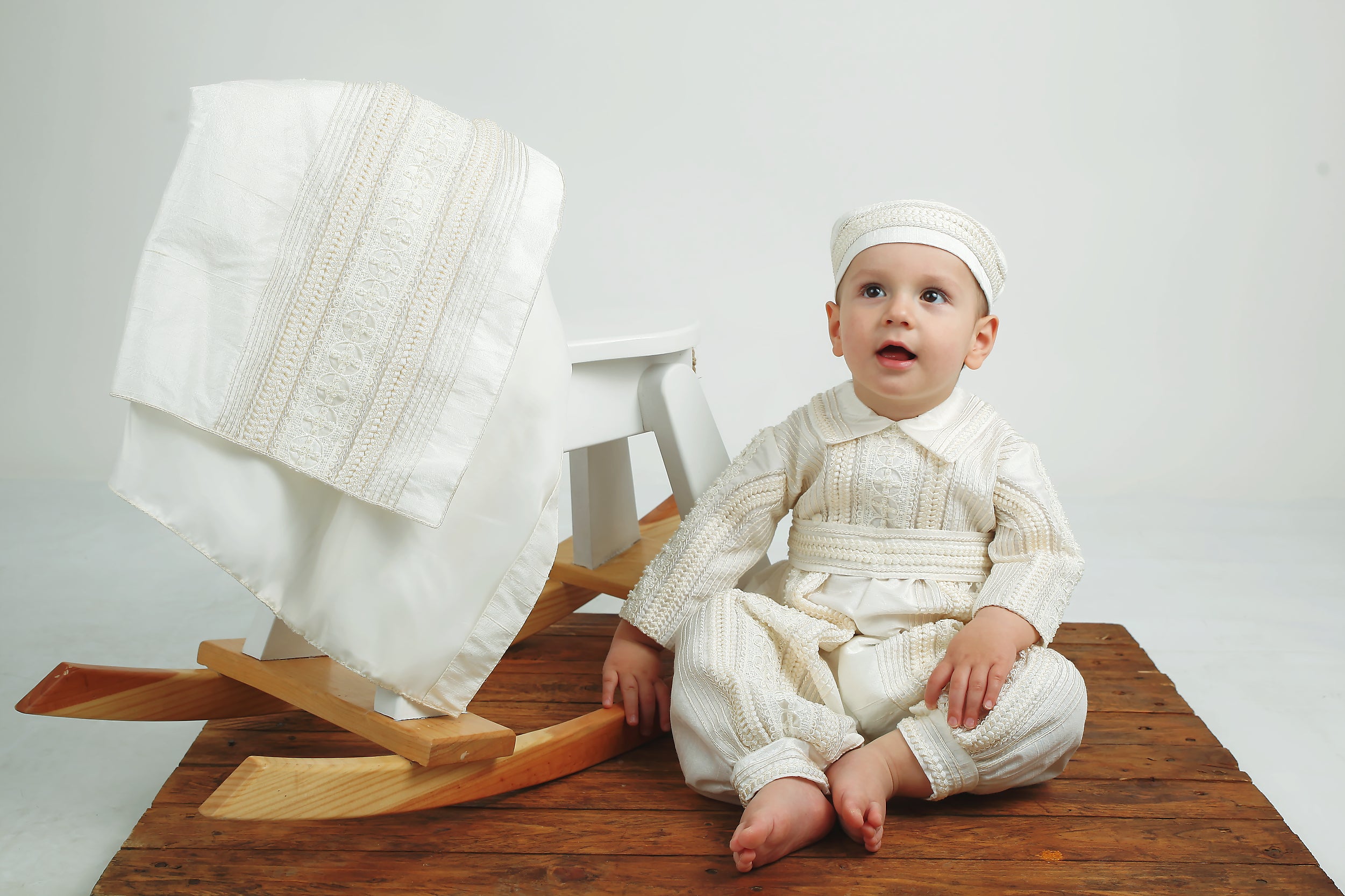Baby boy posing in the Gabriel Dupioni silk romper featuring hand‑finished pearl and lace detailing, detachable skirt draped over rocking horse.