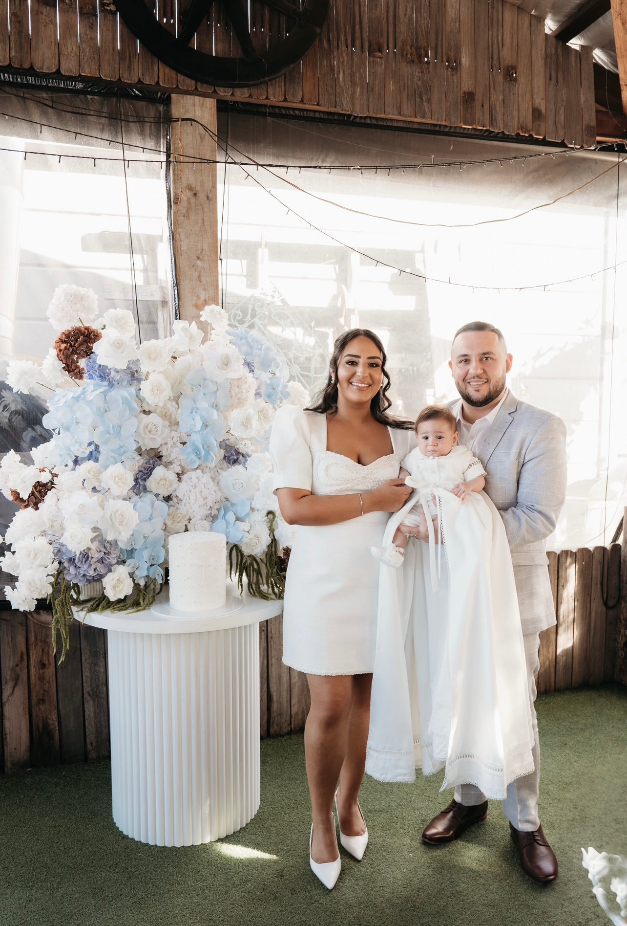 Family moment with mum and dad standing together while baby wears the My Blessing Georgie christening romper, the Mikado fabric giving a classic, formal look.