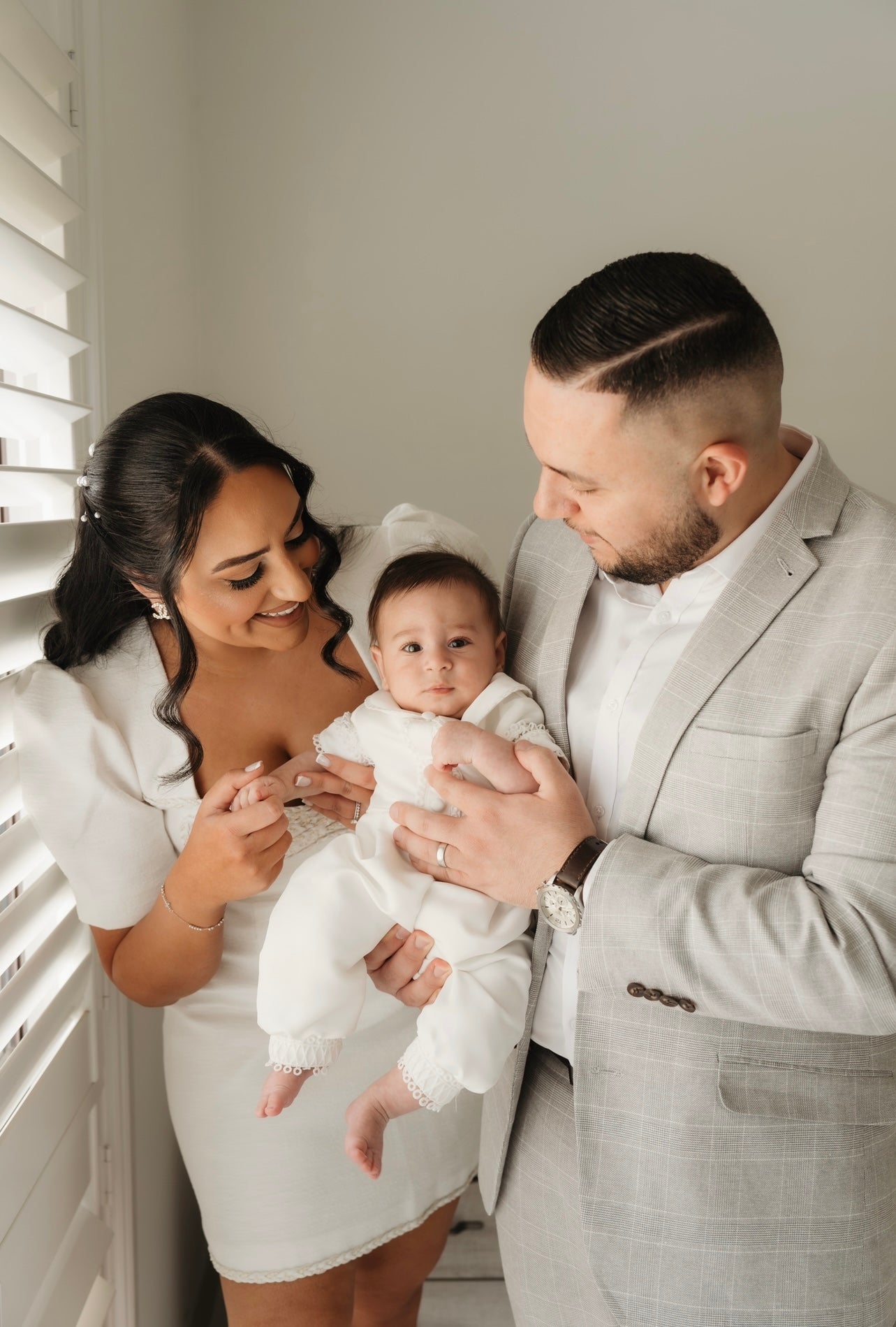 Baby in the My Blessing Georgie christening romper held between mum and dad, the structured Mikado fabric adding elegance to the family portrait.