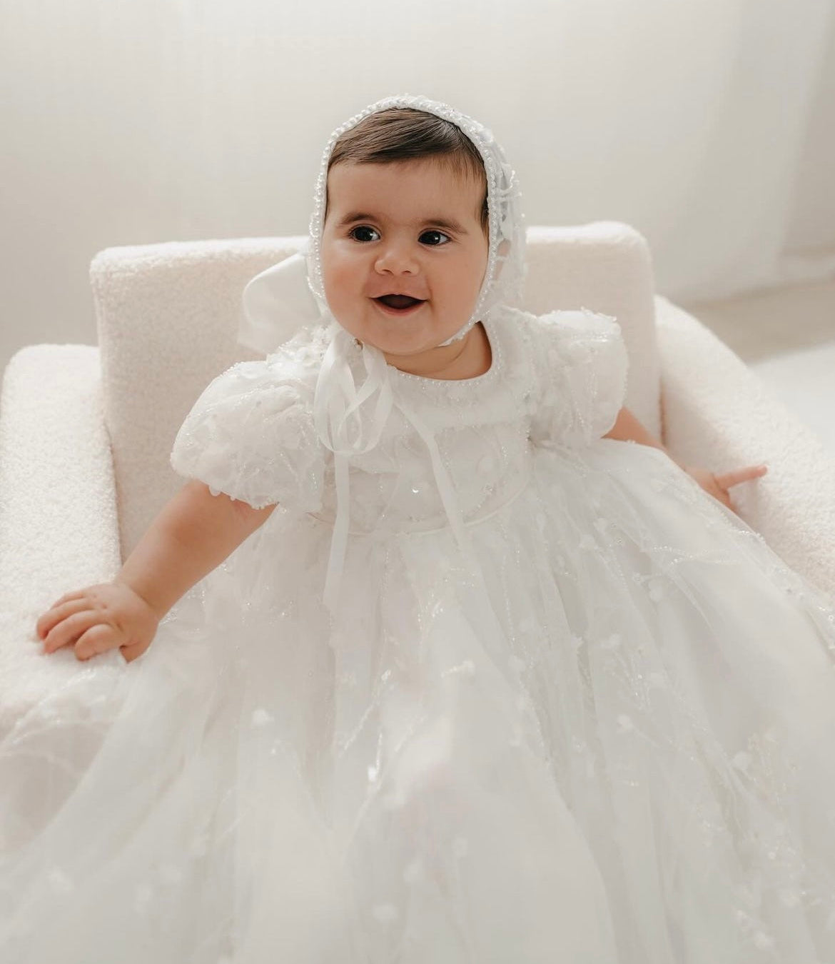 Baby wearing My Blessing Shamiran ivory christening gown while sitting, showing the lace bodice, flowing satin skirt, and matching accessories.
