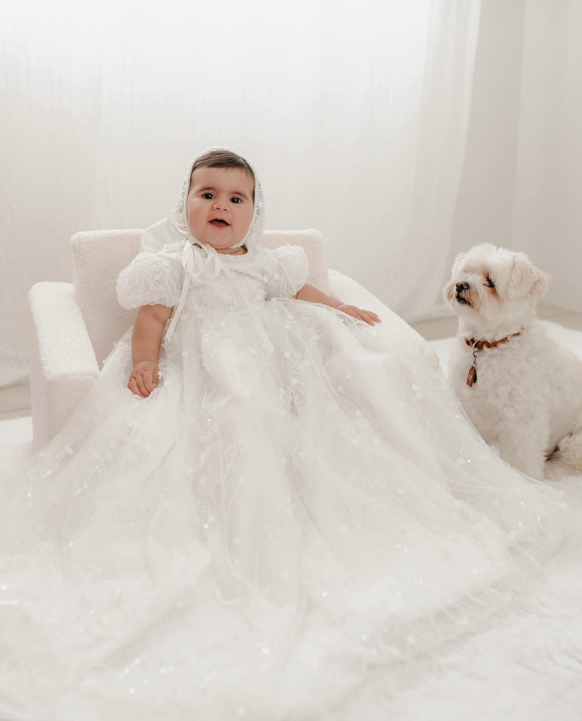 Baby seated showing the front of the My Blessing Shamiran christening gown, highlighting the lace‑embroidered bodice, sleeves, and flowing satin skirt .