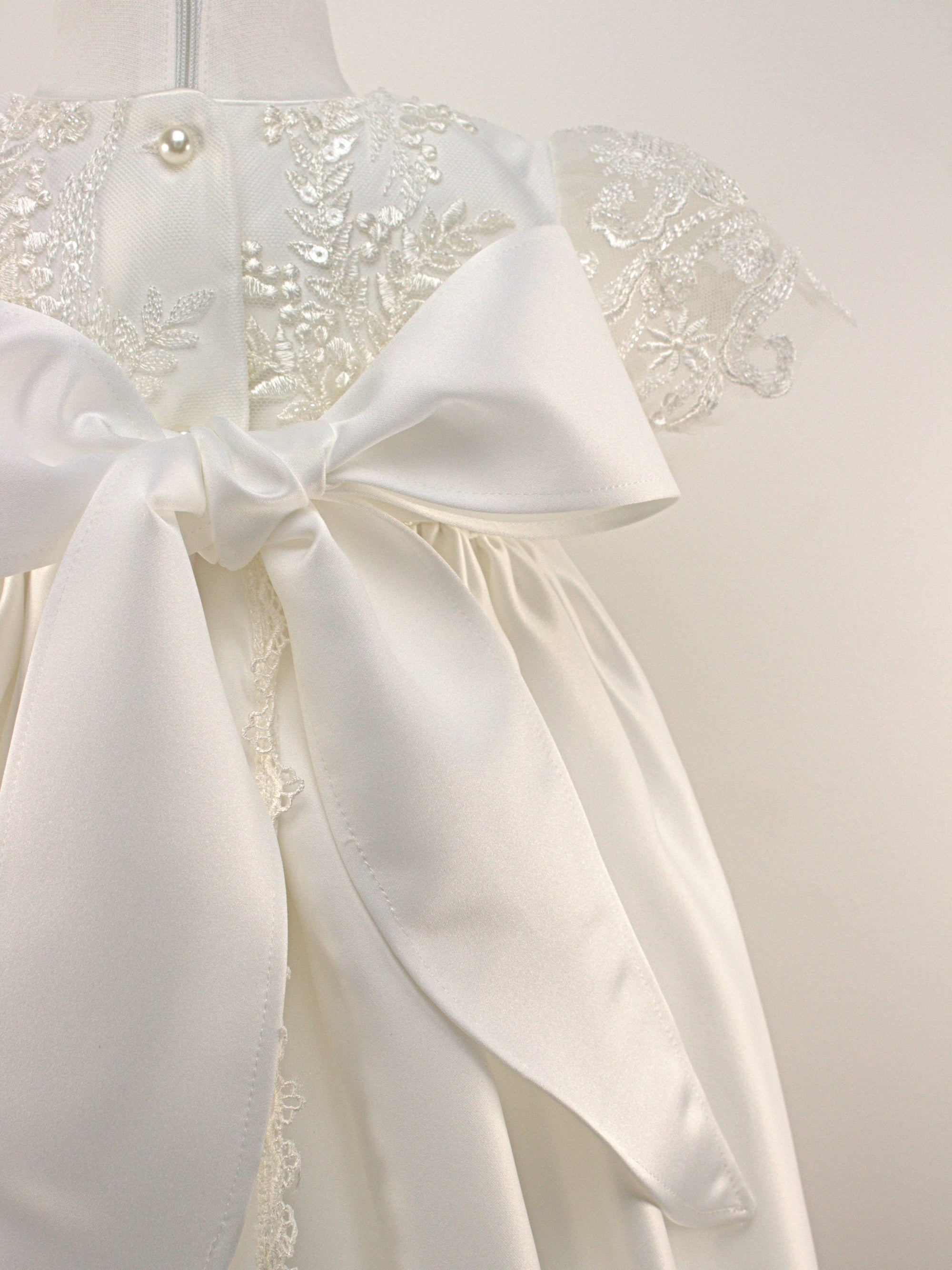 Ivory christening gown on a mannequin featuring detailed floral lace, bead embroidery on the bodice and sleeves, and a softly gathered satin skirt.