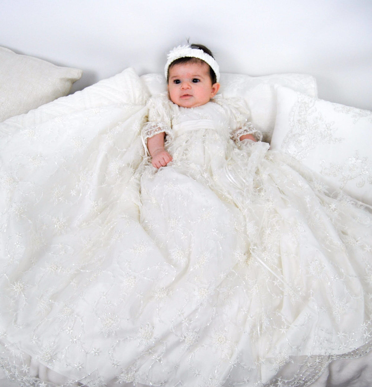 baby girl wearing ivory full‑length My Blessing Devotion christening gown featuring a European floral lace overlay with delicate glass seed bead detailing.