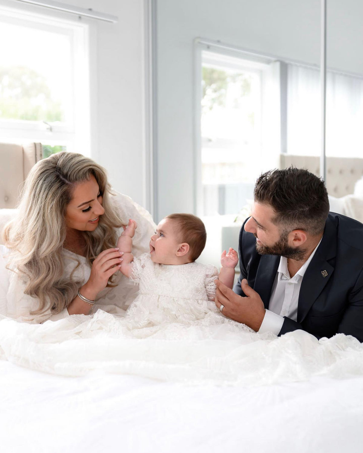 Parents holding their baby girl wearing the My Blessing Elora ivory christening gown, with lace embellished by sequins, glass seed beads, and Swarovski crystal beadwork.