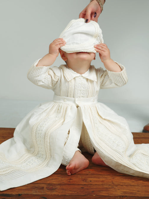 Baby boy playing with hat while wearing the My Blessing Gabriel Dupioni silk romper, the pearl detailing catching the light softly.