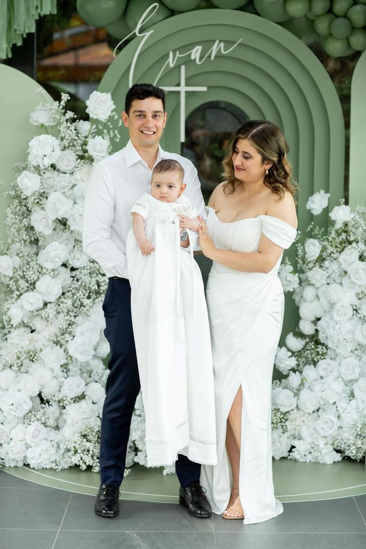 Family photo with mum and dad holding their baby wearing the My Blessing Georgie Mikado romper, the smooth fabric glowing softly in the natural light.