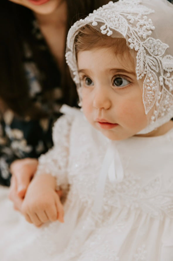 My Blessing Gianna christening gowns on baby girl highlighting front bodice showing intricate lace embroidery embellished with glass seed beads and pearls. Baby girl wearing matching bonnet.