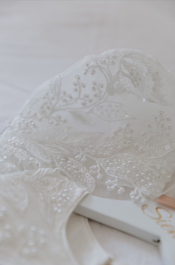 Flat‑lay display of the My Blessing Gianna ivory christening set bonnet showing the embroidered lace with pearl and glass seed bead detailing.