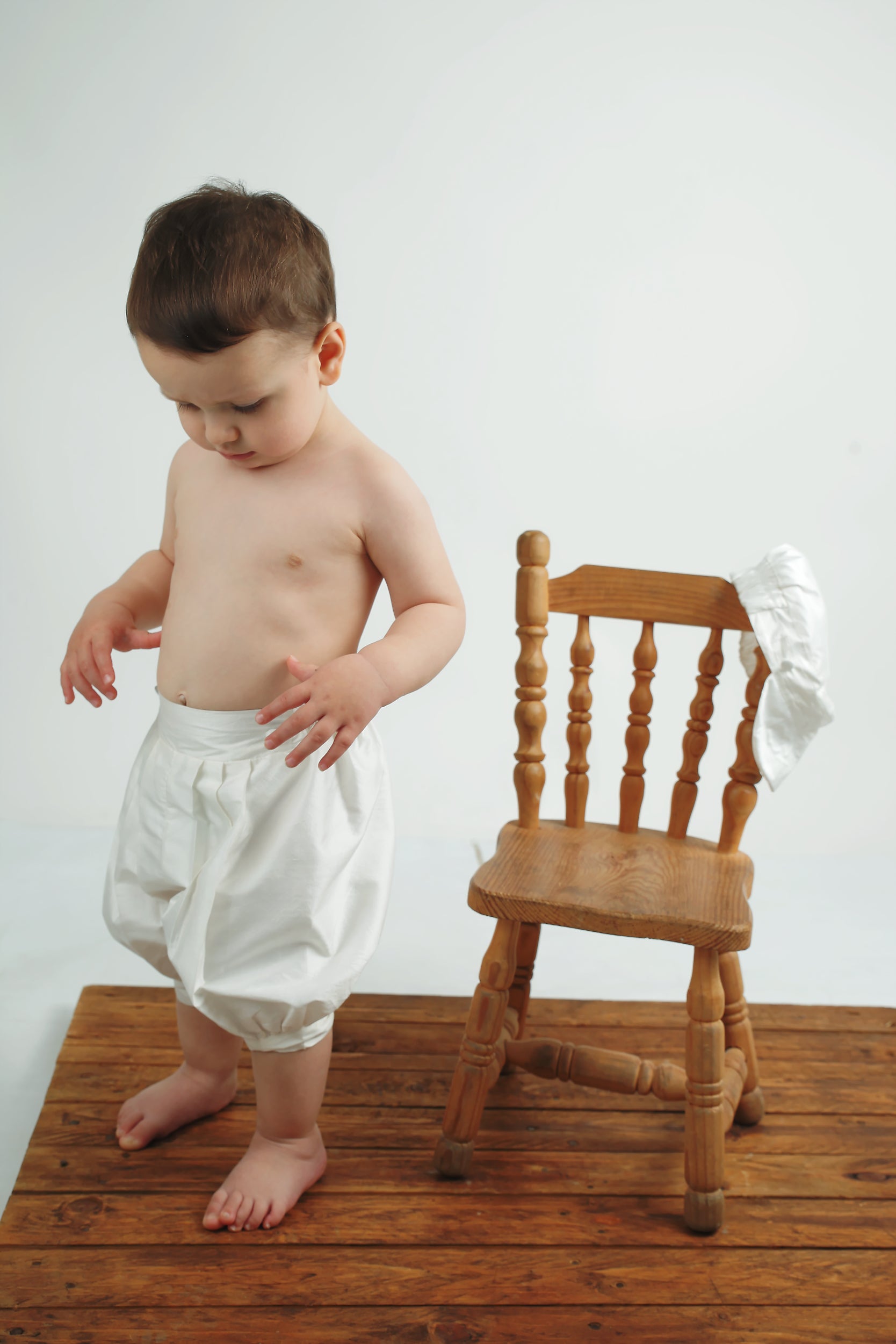 Baby boy posing in the My Blessing Lemuel Dupioni silk pants featuring refined, vintage‑inspired styling with cap draped over chair.