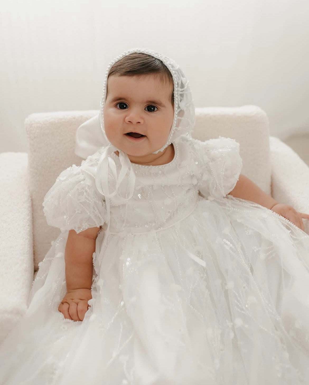 Baby girl seated softly while wearing the My Blessing Shamiran Gown, featuring delicate lace embroidery and a flowing satin skirt.