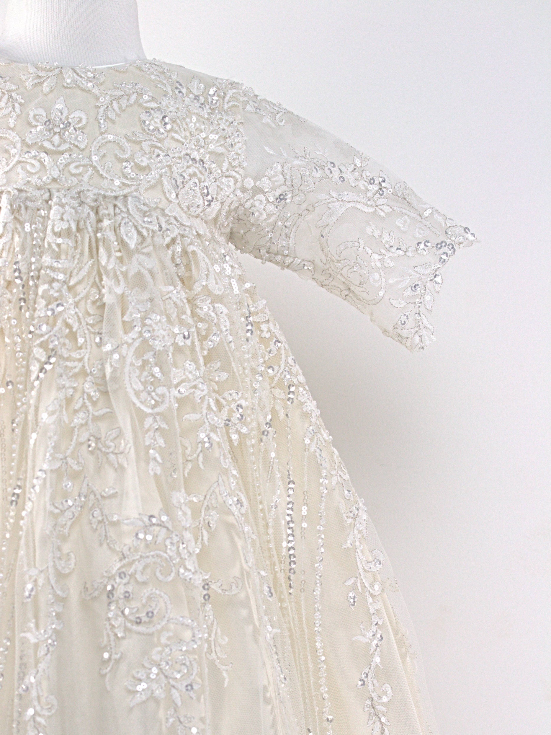 A detail image of the My Blessing Wholesome ivory full-length baby gown with silver sequins and lacework, featuring mid-length sleeves and a large silk bow at the back.