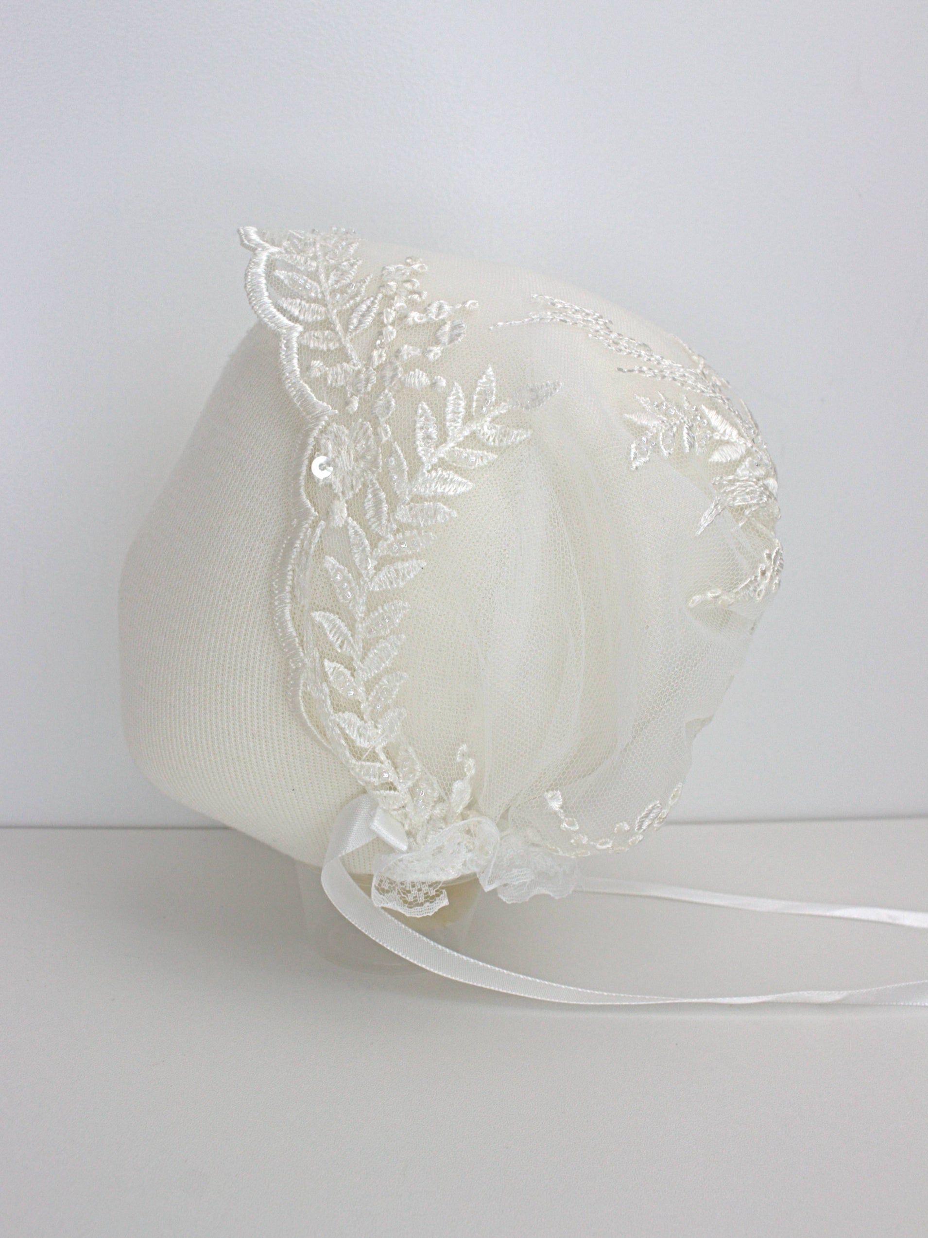 Ivory christening bonnet with floral lace embroidery and soft satin ties, displayed on a mannequin.