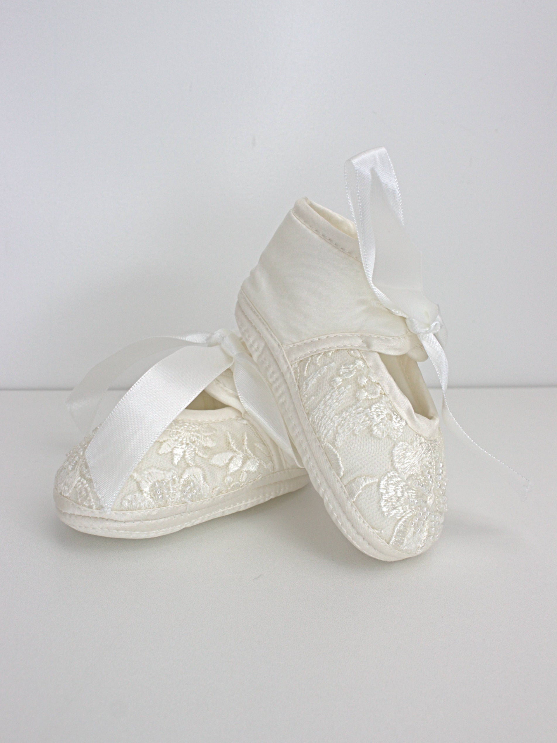 Ivory baby christening shoes with satin finish and lace embellishment, shown as a matching pair.